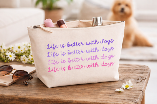 Beige tote bag with 'Life is better with dogs' text, sunglasses, and flowers on a wooden surface.