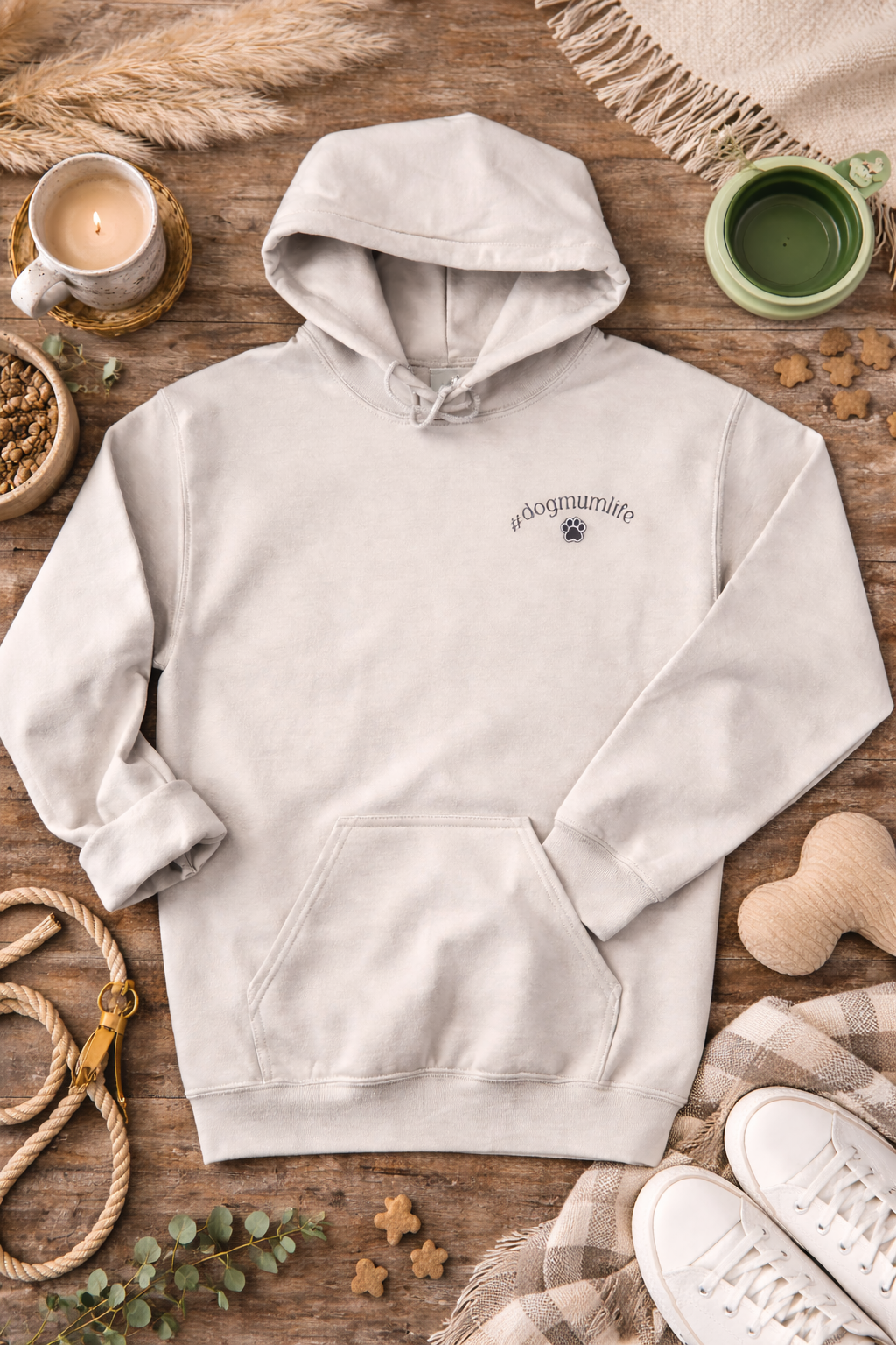 Cream hoodie with a small logo on a wooden surface with a cup, candle, and dog toys.