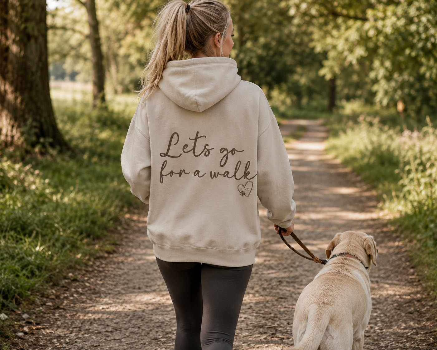 Woman wearing a sand "Let's Go for a Walk" hoodie walking a Labrador in a country park. Dog lover hoodie featuring a bold back design, ideal for dog walking, pet owners, and outdoor lifestyle clothing from That Woofin Dog. 🐶🍂