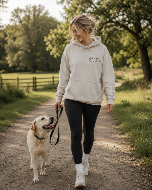 Woman wearing a sand "Let's Go for a Walk" hoodie walking a Labrador in a country park. Dog lover hoodie featuring a small chest design, ideal for dog walking, pet owners, and outdoor lifestyle clothing from That Woofin Dog. 🐶🍂